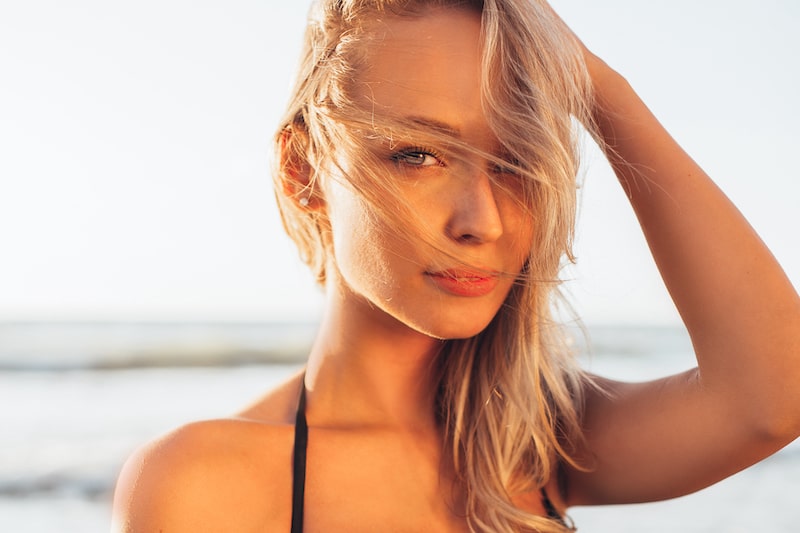 Beautiful blonde woman with tan skin looks at camera with beach in background.